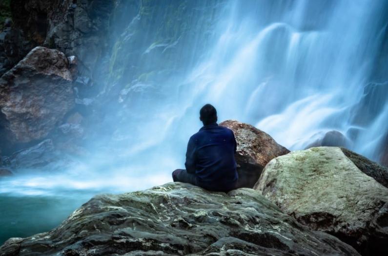 Man sitting at rock in front of waterfall white water stream falling from mountains.
