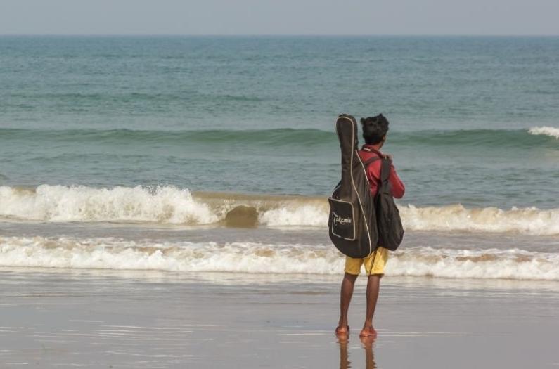 A boy stands with his guitar facing the sea on the Rushikonda beach in a tranquil, peaceful moment.