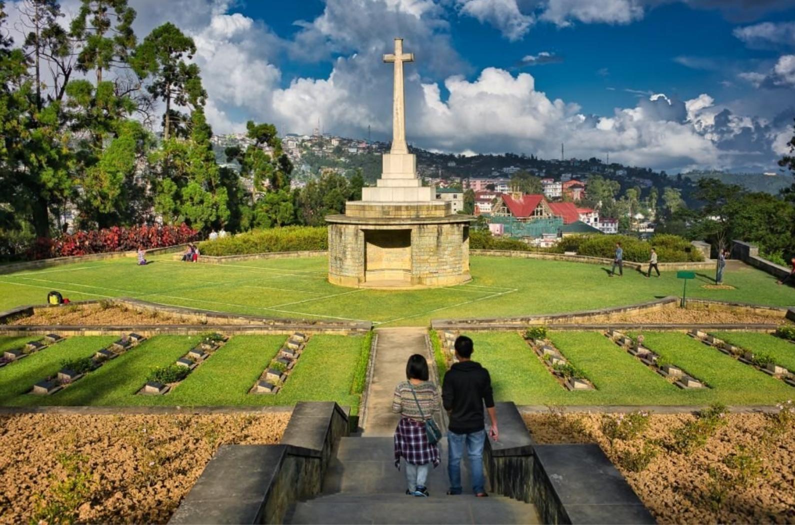 Two people in front of the big white cross of Kohima War Cemetery, while other people take photos and admire the cemetery on a sunny day, in Nagaland region, in Seven Sisters of India.