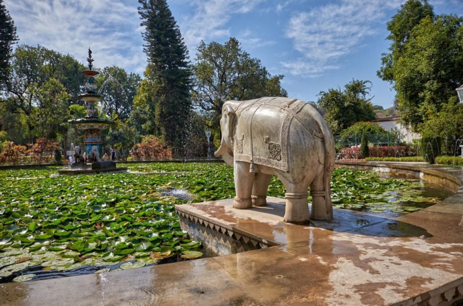 Elephant statue facing into a large fountain. Lilly pads in the water. Blue sky.