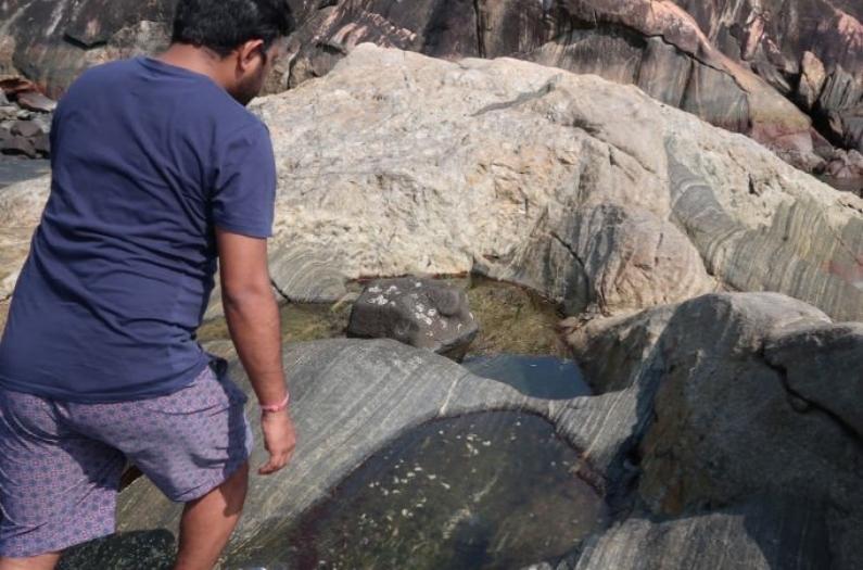 Indian man enjoying sunshine and warmth whilst walking on boulders and looking at rock pools at Colomb Beach.